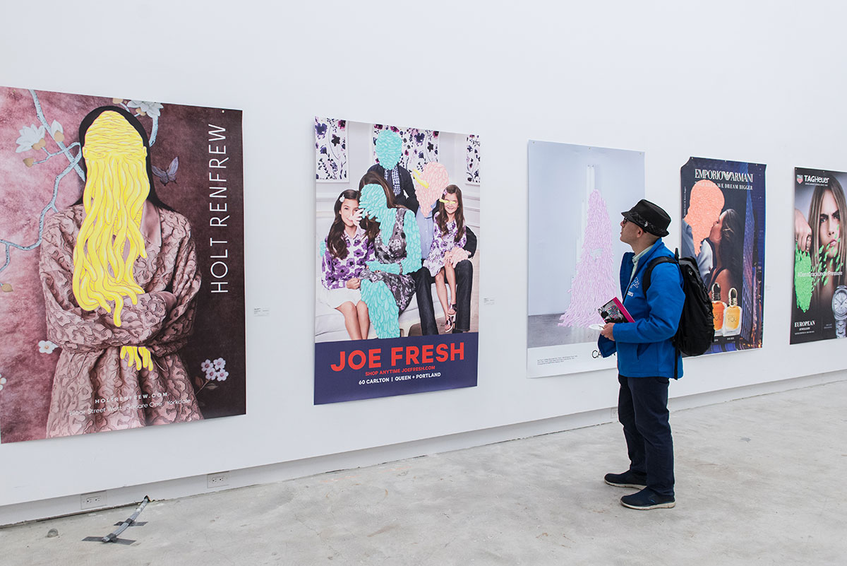 A person in a gallery viewing wall-based artwork featuring large advertisements altered with hand-painted elements.