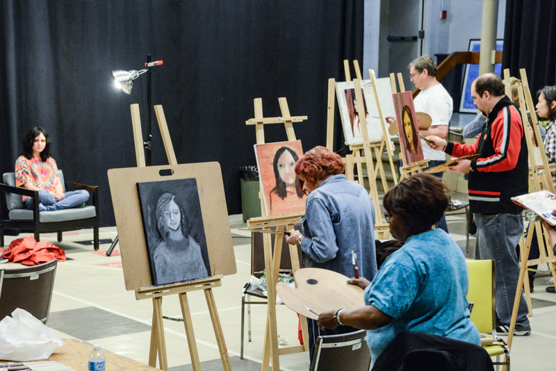 A group of people at easles, painting a model in a chair at the front of the room.