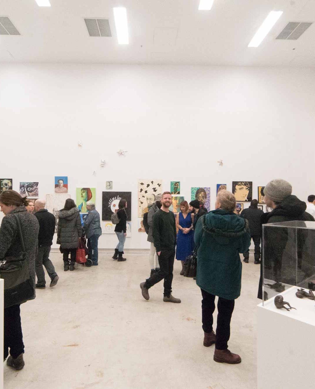 A large group of people fill a gallery space with plinths and artwork on walls at an opening reception.