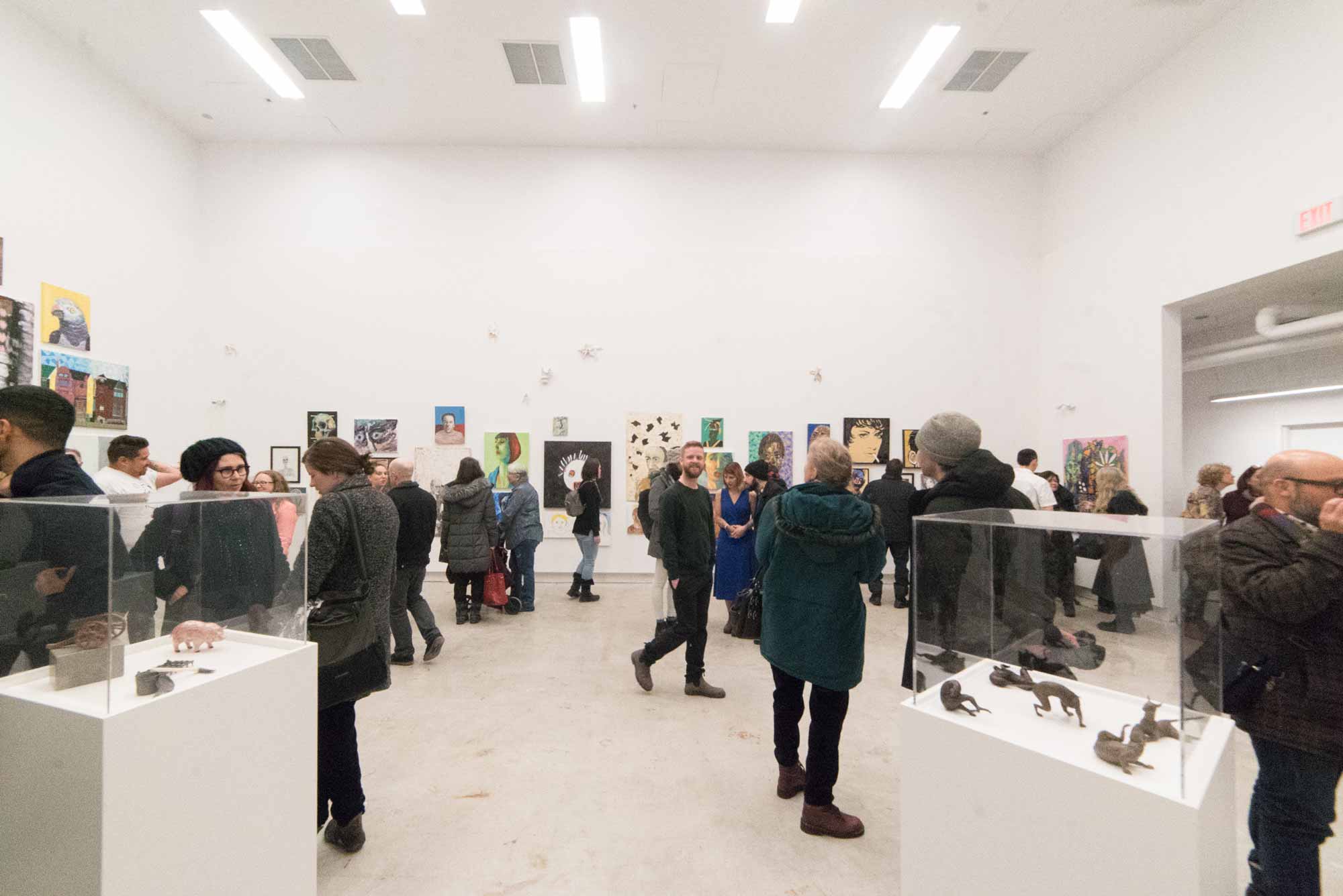 A large group of people fill a gallery space with plinths and artwork on walls at an opening reception.