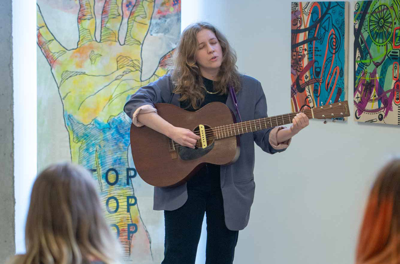 A person standing, singing and playing guitar in front of other people sitting in chairs. There is art on walls in the background.