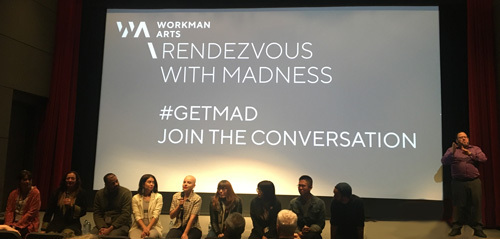 9 people sit on a stage edge, one holding a microphone. An ASL interpreter is behind & to the right. A screen behind them says Workman Arts Rendezvous with Madness #GetMad Join the Conversation