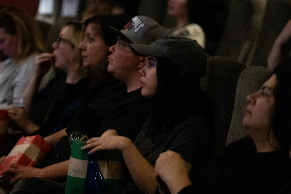 Audience members in their seats, looking at the screen. Two of them are holding popcorn.