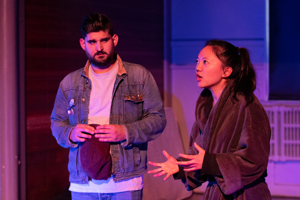A man with a beard in a jean jacket hold a hat in his hands while watching a woman in a bathrobe with her hair in a ponytail speak. She is gesturing with her hands.