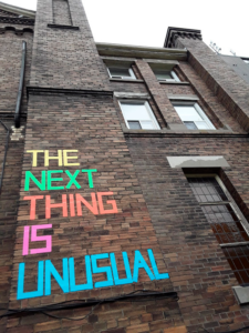 A brick building with the words "The next thing is unusual" written on it colourful duct tape.