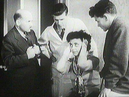 A black and white film still from the 1951 film “Breakdown” depicting a woman being examined by three men.