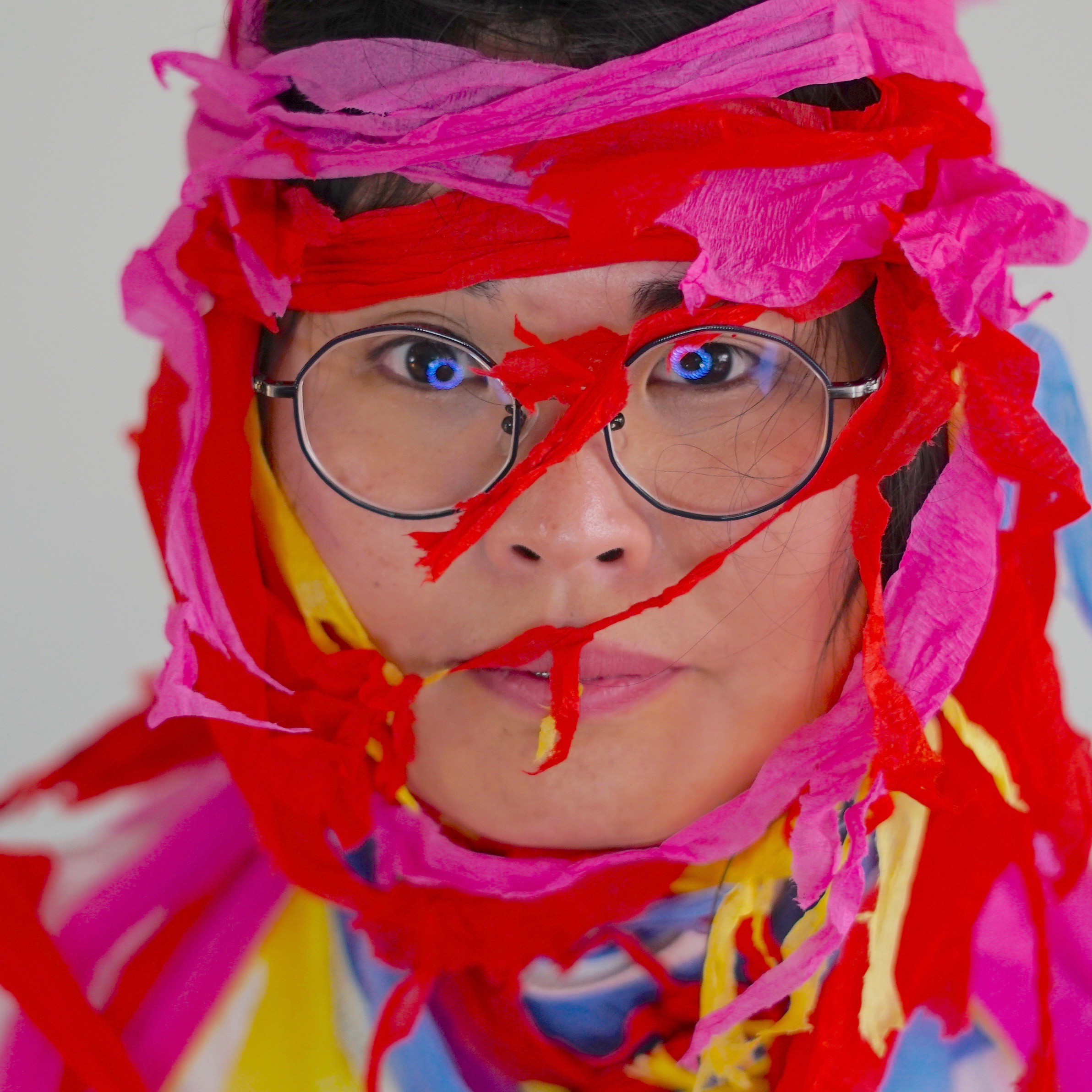 Headshot of a person facing the camera with vividly colored stretchy paper strips wrapped around their head.