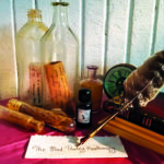 A photographic still life image with an ink bottle, books, a round analog clock with Roman numerals, large transparent bottles containing handwritten messages on yellowed paper, and a quill pen spelling out “Mad Poetry Apothecary” on a piece of paper.
