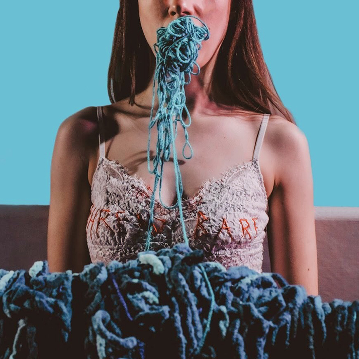 A cropped photo of a person in a lacy top with the word “threadbare” embroidered across the chest. Chunky blue-green yarn streams out of their mouth and fills the foreground of the image.