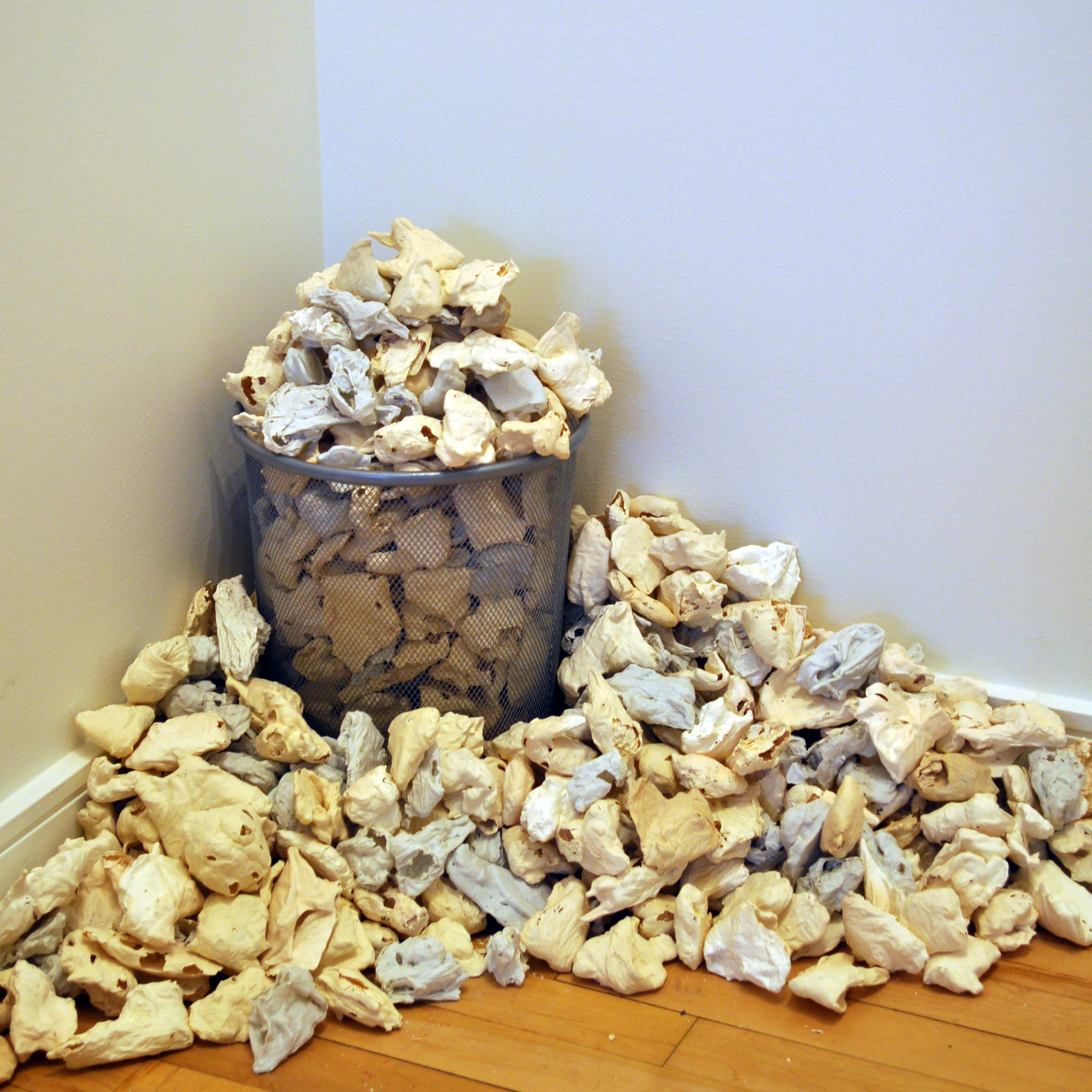 A photograph depicting a wire wastebasket in the corner of a room, overflowing with crumpled tissues covered in a smooth, hard yellowish or grayish substance.