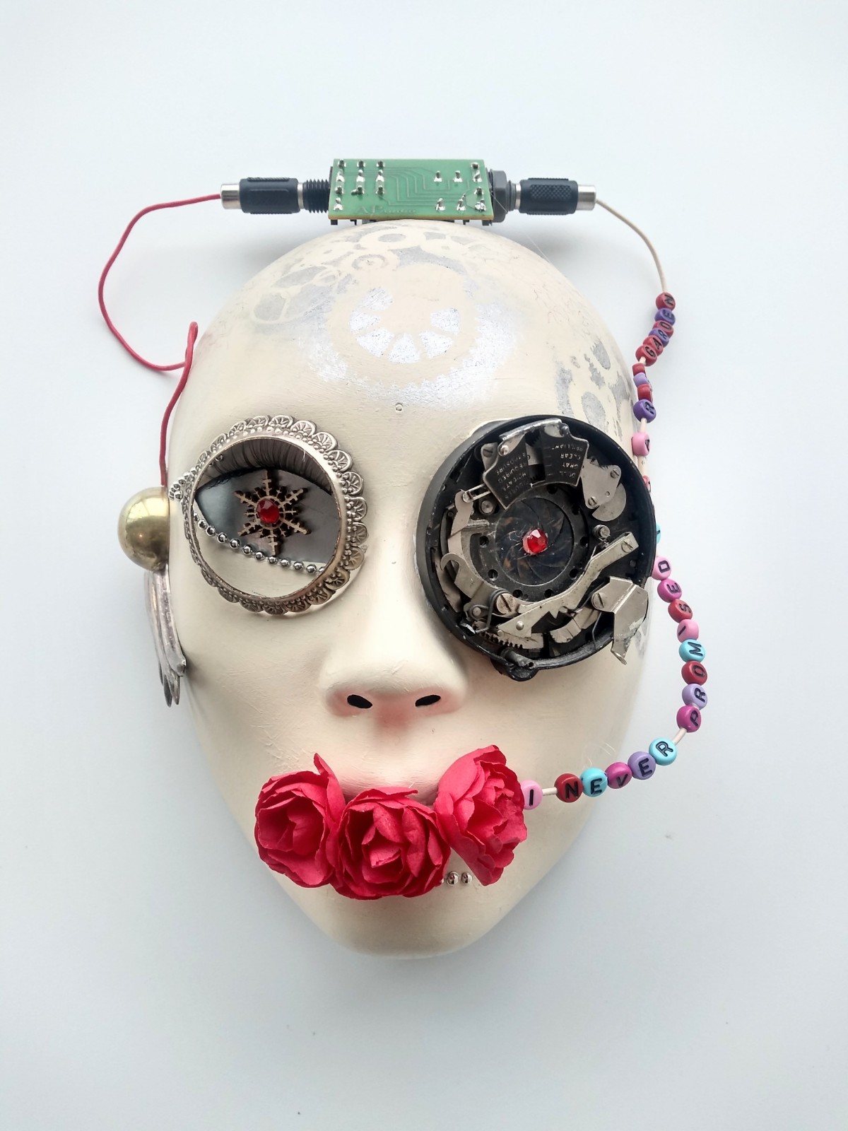 A 3-D white mask with steampunk style fixtures. Three red roses coming out of its mouth and beads on the side of its head that read "I never promised you a rose garden"