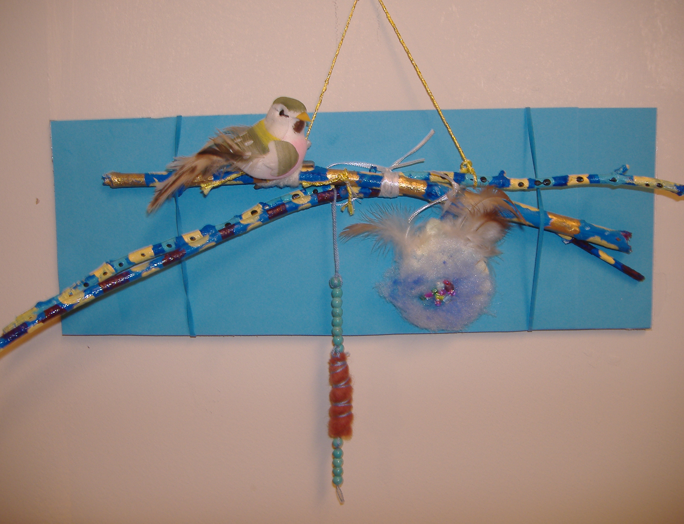 A blue rectangle backing holds a painted branch with a bird and beads hung on a string.