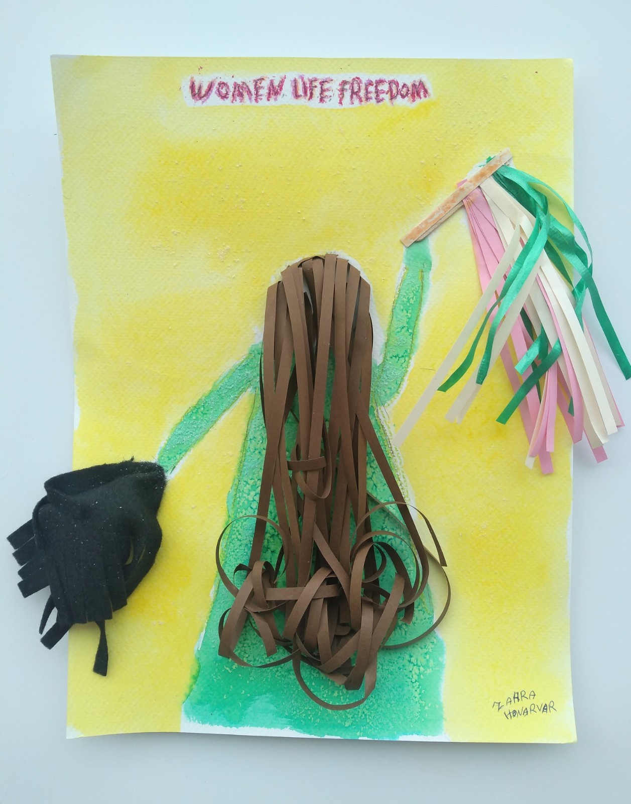 A yellow background piece with text at the top "Women Life Freedom". The back of a person in green holds pom pom per hand and brown hair. Both the pompoms and hair are 3-D.