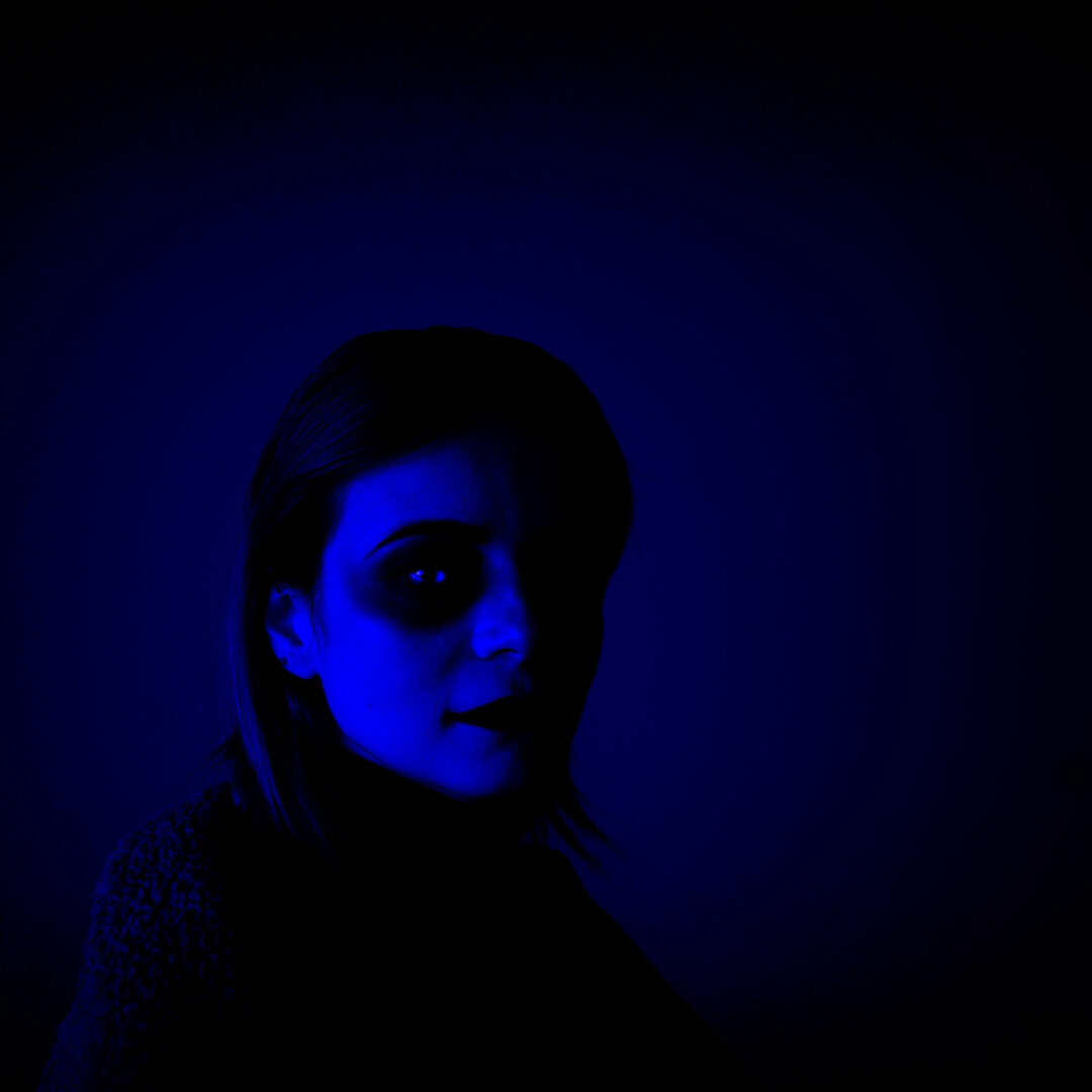 A person with shoulder length hair faces the camera with dark circle around the eye and a blue filter in the dark portrait.