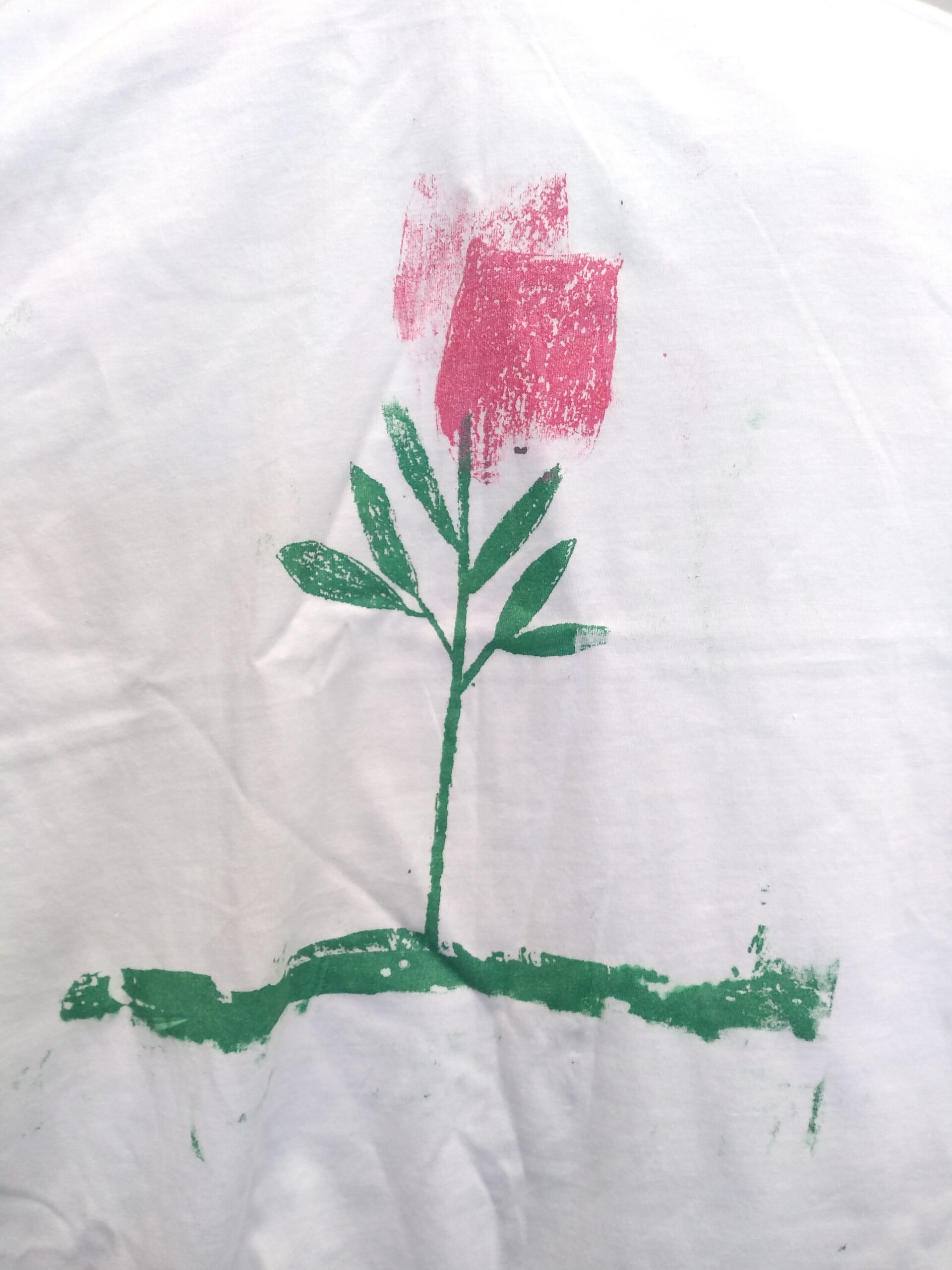 Red rose with green stem and leaves on a green coloured ground on white shirt.