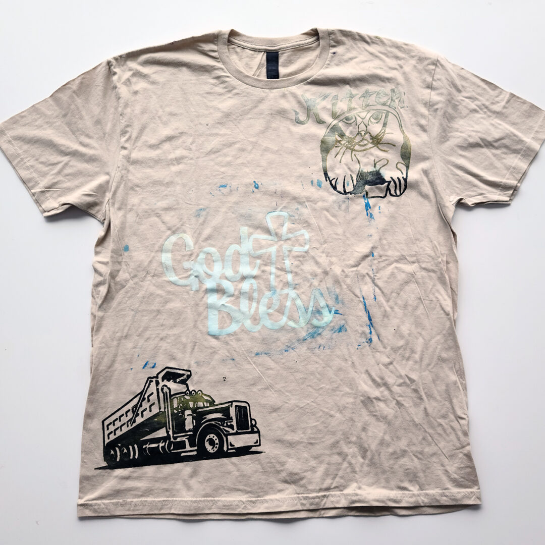 Beige t shirt screenprinted with an illustration of a cat, the words 'God Bless' and a truck.