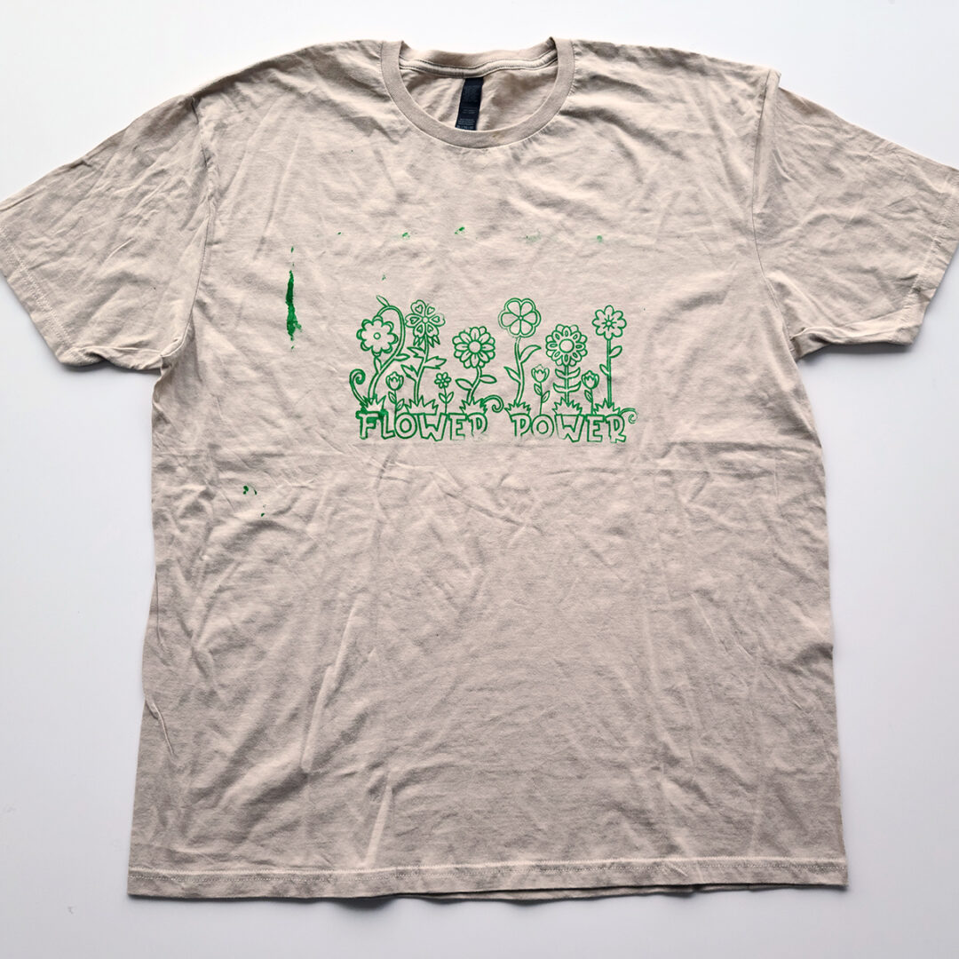 Beige t shirt screenprinted with six flowers with the words 'Flower Power' below in green.